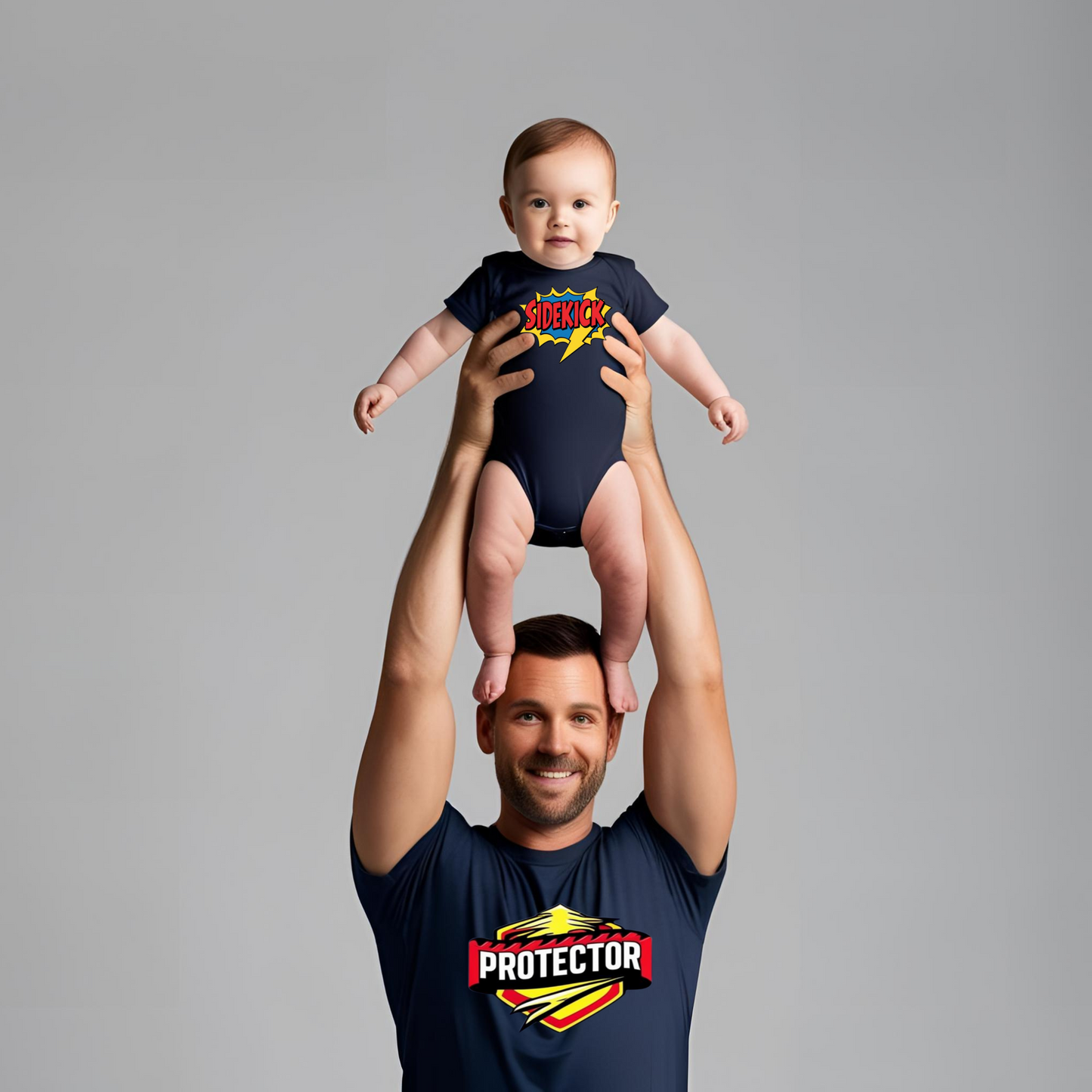 Dad lifting baby in superhero pose, wearing matching navy “Protector” T-shirt and “Sidekick” onesie, smiling together in playful moment.