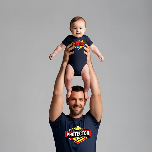 Dad lifting baby in superhero pose, wearing matching navy “Protector” T-shirt and “Sidekick” onesie, smiling together in playful moment.